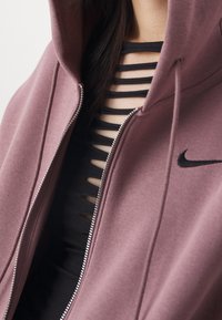 Rosafärgad hoodie med dragkedja och snoddar, kombinerad med en svart åtsittande topp med horisontella urtag. Nike-logotyp synlig.