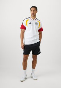 Polo branco com detalhes em vermelho, amarelo e preto, apresentando o emblema da seleção de futebol da Alemanha. Acompanhado de calções pretos e ténis brancos.