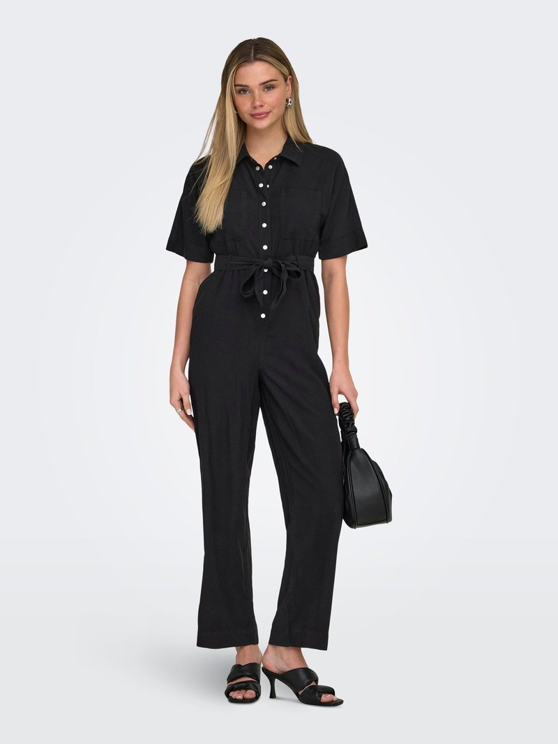 Femme aux longs cheveux blonds se tient debout, portant une combinaison noire boutonnée avec ceinture, tenant un sac à main noir et portant des sandales à talons noirs.