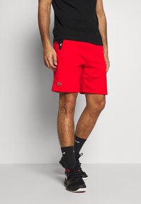 Röda idrottsshorts med en slät yta och resårmidja, med sidofickor och en liten kontrasterande logotyp.