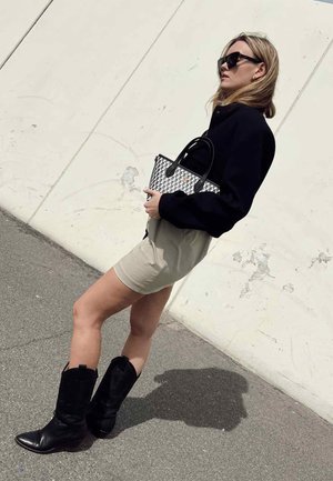 Zwarte oversized trui, beige shorts, zwarte kniehoge laarzen; houdt een geruite handtas met zwarte handvatten vast tegen een lichte muur.