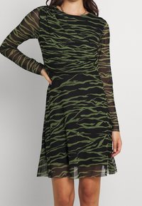 Musta mekko, jossa on pitkät läpinäkyvät hihat, oliivinvihreä tiikeriraidallinen kuosi, muotoiltu yläosa ja levenevä hame, jossa on sivusilto.