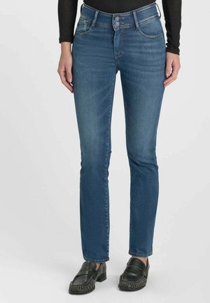 Femme portant un jean droit bleu moyen avec une taille à double bouton et des mocassins en cuir noir, debout devant un fond uni.