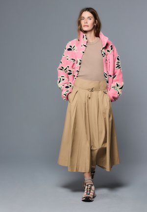 Frau trägt eine pink gemusterte Jacke über einem beigen Top, beige weit ausgestellte Hose, Socken und Turnschuhe, steht vor einem grauen Hintergrund.