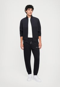 Herre treningstøy med en svart jakke med glidelås og ensfarget design, samt matchende bukser, begge laget av mykt stoff, kombinert med hvite joggesko.