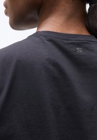 Κοντινό πλάνο του λαιμού και της άνω πλάτης ενός ατόμου που φοράει σκούρο γκρι πουκάμισο με ένα μικρό ανακλαστικό λογότυπο "S" κοντά στον γιακά.