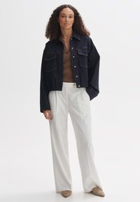 Veste en denim foncé avec deux poches avant, associée à un haut en maille marron et un pantalon large crème. Le modèle porte des chaussures beiges.