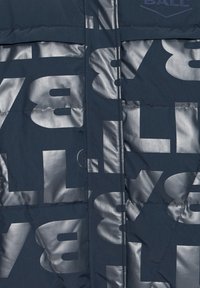 Chaqueta acolchada en azul marino oscuro con grandes gráficos metálicos plateados. Presenta textura acolchada, cremallera frontal y detalle de logo de tela.