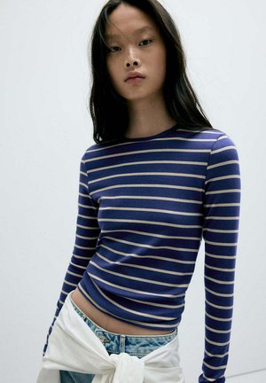 Jeune femme aux longs cheveux foncés portant une chemise à manches longues rayée bleu et blanc et un jean en denim clair avec un pull blanc noué autour de la taille.