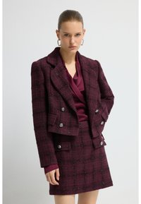 Veste en tweed bordeaux avec un design croisé, des accents de boutons argentés et une jupe à carreaux assortie ; superposée à un chemisier violet profond.