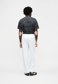 Μαύρο πουκάμισο με κοντά μανίκια και σχέδιο, με άνετη εφαρμογή, συνδυασμένο με ανοιχτό γκρι φόρμα. Το μοντέλο φοράει μαύρα αθλητικά παπούτσια με λευκές λεπτομέρειες.