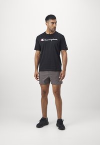 Schwarzes Baumwoll-T-Shirt mit weißem "Champion"-Logo, kombiniert mit grauen Shorts und schwarzen Turnschuhen, stehend vor einem neutralen Hintergrund.