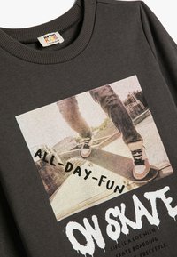 Ciemnoszary sweatshirt z grafiką na przodzie przedstawiającą skateboardzistę oraz tekstem "WSZYSTKIE DNI ZABAWY" i "NA DESCE" w wyraźnych, białych czcionkach.