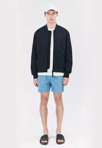Marine bomberjack, lichtgewicht stof, rits aan de voorkant, zijzakken; gecombineerd met een wit T-shirt, blauwe shorts en zwarte instap sandalen.