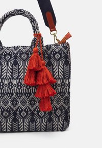Textilní taška s námořnickým a krémovým geometrickým vzorem, s oranžovými třásněmi a koženými detaily. Obsahuje ramenní popruh a zip.