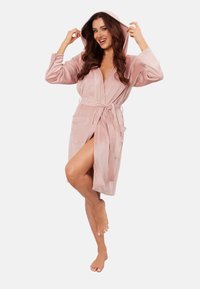 Jasnoróżowy, kapturowy szlafrok wykonany z miękkiego materiału, z długimi rękawami, paskiem w talii i dwiema przednimi kieszeniami. Prezentowany z nogami lekko skrzyżowanymi.