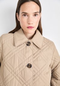 Beige quilted jacket with a large collar, featuring a dark button and diagonal stitching patterns. Smooth fabric texture, designed for cool weather.