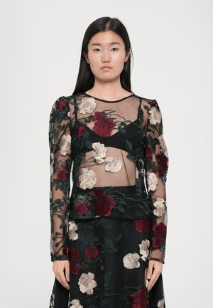 Vestido preto transparente com mangas longas, apresentando intrincados bordados florais em vermelho e bege. O tecido transparente sobrepõe seções sólidas em preto.
