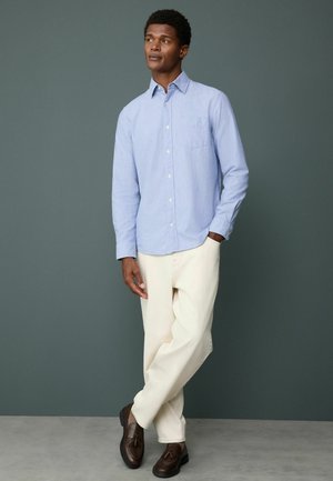 Helles blaue Langarmhemd mit Knopfdown-Kragen, beigen Hosen und braunen Schuhen, stehend vor einem dunkelgrünen Hintergrund. Minimale Taschendetails.