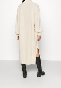 Abito beige lavorato a maglia con maniche lunghe, colletto alto e spacco laterale; abbinato a stivali neri al ginocchio. Design rilassato con texture.