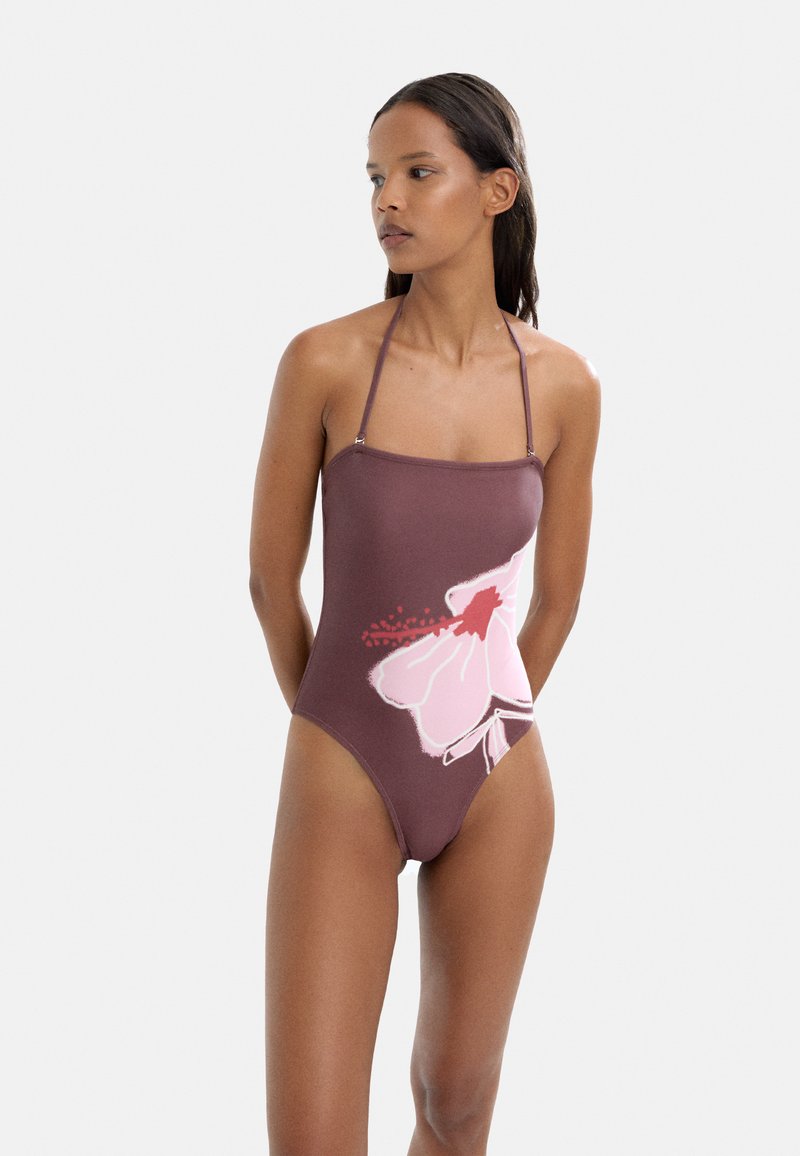 Jednoczęściowy strój kąpielowy w ciemnym brązie z dużym różowym wzorem kwiatu po lewej stronie. Posiada cienkie ramiączka i wysoki krój nogawki.