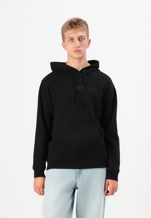 Sort hættetrøje med en kængurulomme og snoretræk, der har et diskret GAP-logo på brystet. Bæres med lyseblå jeans.