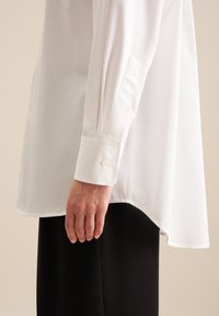 Wit overhemd met lange mouwen, voorzien van een high-low zoom, knoopmanchetten en een gladde textuur. Draag het met zwarte broek voor een casual uitstraling.