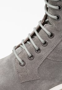 Harmaa mokkasiiniboot, jossa on pyöreät hopeiset niitit, harmaat nauhat ja teksturoitu pinta. Mukana on myös ommeltu yksityiskohta sivussa.