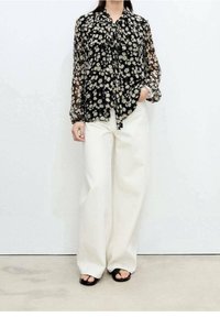 Blouse noire transparente à fleurs avec manches longues et un lien au col, associée à un pantalon large blanc et des sandales noires.