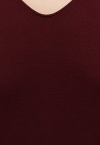 Gros plan d'une personne portant un pull en maille rouge foncé à col en V sur une peau claire.