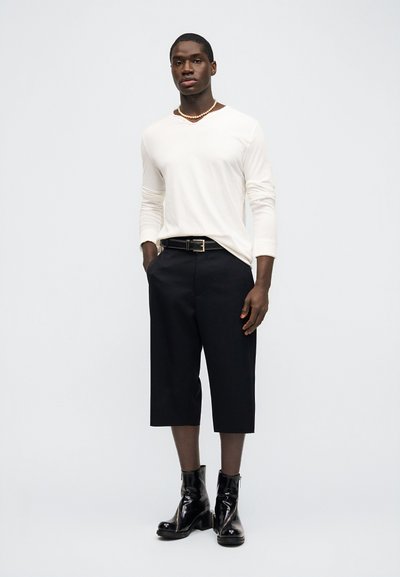 Άνδρας που φοράει λευκό πουκάμισο με μακριά μανίκια, μαύρο σορτς μέχρι το γόνατο, μαύρη ζώνη, κολιέ με πέρλες και μαύρα μποτάκια αστραγάλου, στέκεται μπροστά σε λευκό φόντο.