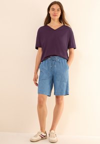 Camiseta de cuello en V morada combinada con pantalones cortos de tiro ajustable en azul claro. Los pantalones cortos cuentan con dos bolsillos frontales y un corte relajado. Usados con zapatillas blancas.