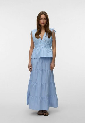 Vrouw draagt een lichtblauw, mouwloos top met V-hals en strikbandjes en een bijpassende gelaagde maxirok met subtiele strepen, staand op een effen achtergrond.