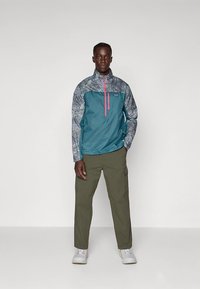 Blågrønn vindjakke med grå mønstrete ermer og rosa glidelås. Kombinert med olivengrønne cargobukser og hvite joggesko. Ensfarget bakgrunn.