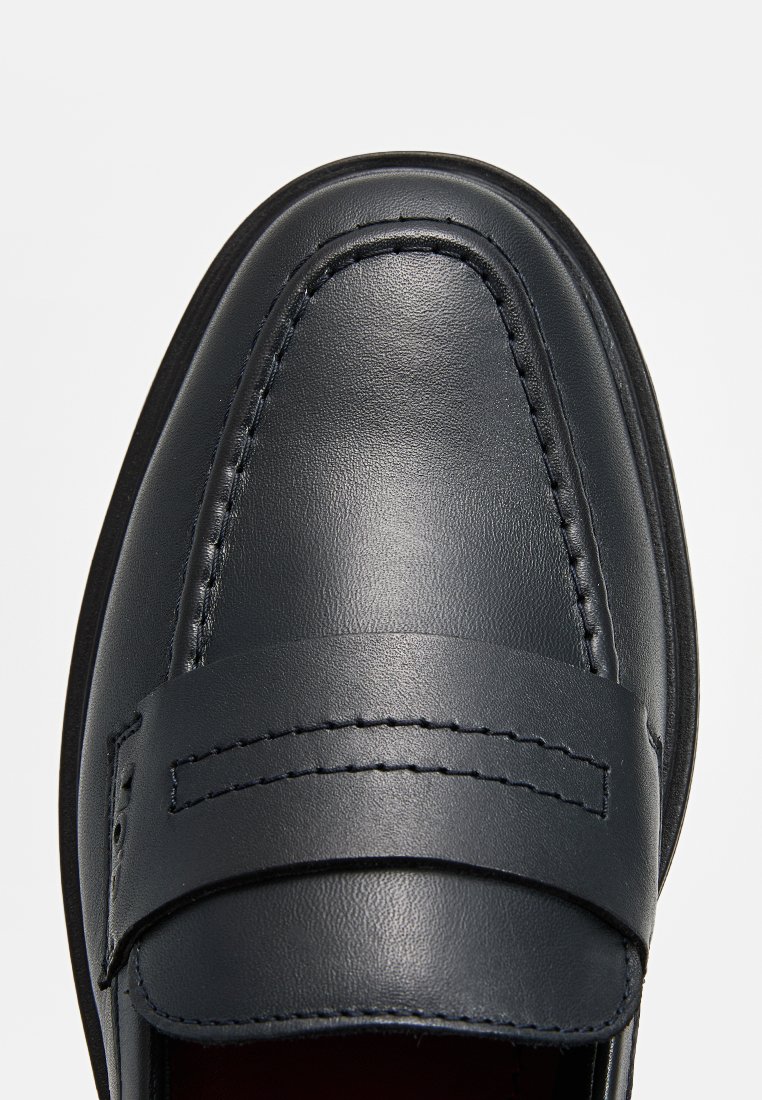 Mocassin en cuir noir avec une texture lisse, doté d'une couture et d'une bande horizontale sur le dessus pour accentuer le style. Bout arrondi.