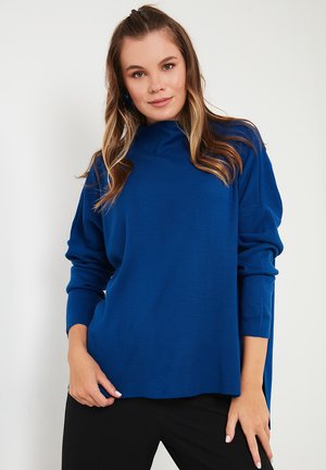 Blauwe oversized pullover met geribbelde textuur, hoge halslijn en dropped shoulders. De stof is zacht, met zijsplitten aan de zoom.