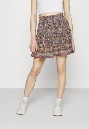Femme portant une jupe à motif floral avec un ourlet à volants, un top court blanc, des chaussettes blanches et des baskets blanches, debout devant un fond uni.