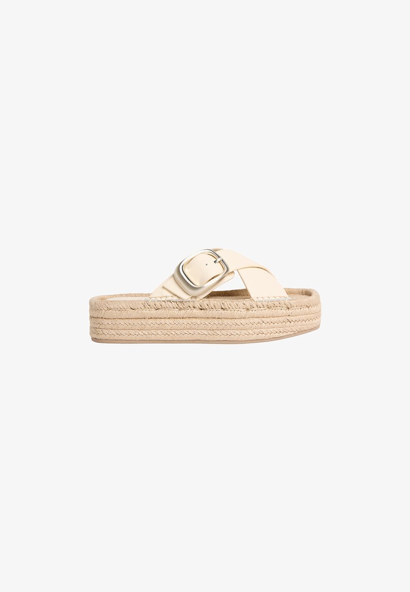 Sandale à plateforme beige avec semelle tressée et larges brides croisées ornées d'une grande boucle métallique sur fond blanc.