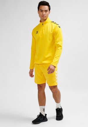 Gelbes Sportoutfit, bestehend aus einem langärmeligen Oberteil mit einem Reißverschluss und passenden Shorts. Besitzt schwarze Akzente an den Seiten und kontrastierende weiße Streifen.