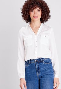 Blusa bianca a maniche lunghe con due tasche sul petto, chiusura frontale con bottoni e dettagli sulle spalle, abbinata a jeans blu a vita alta.