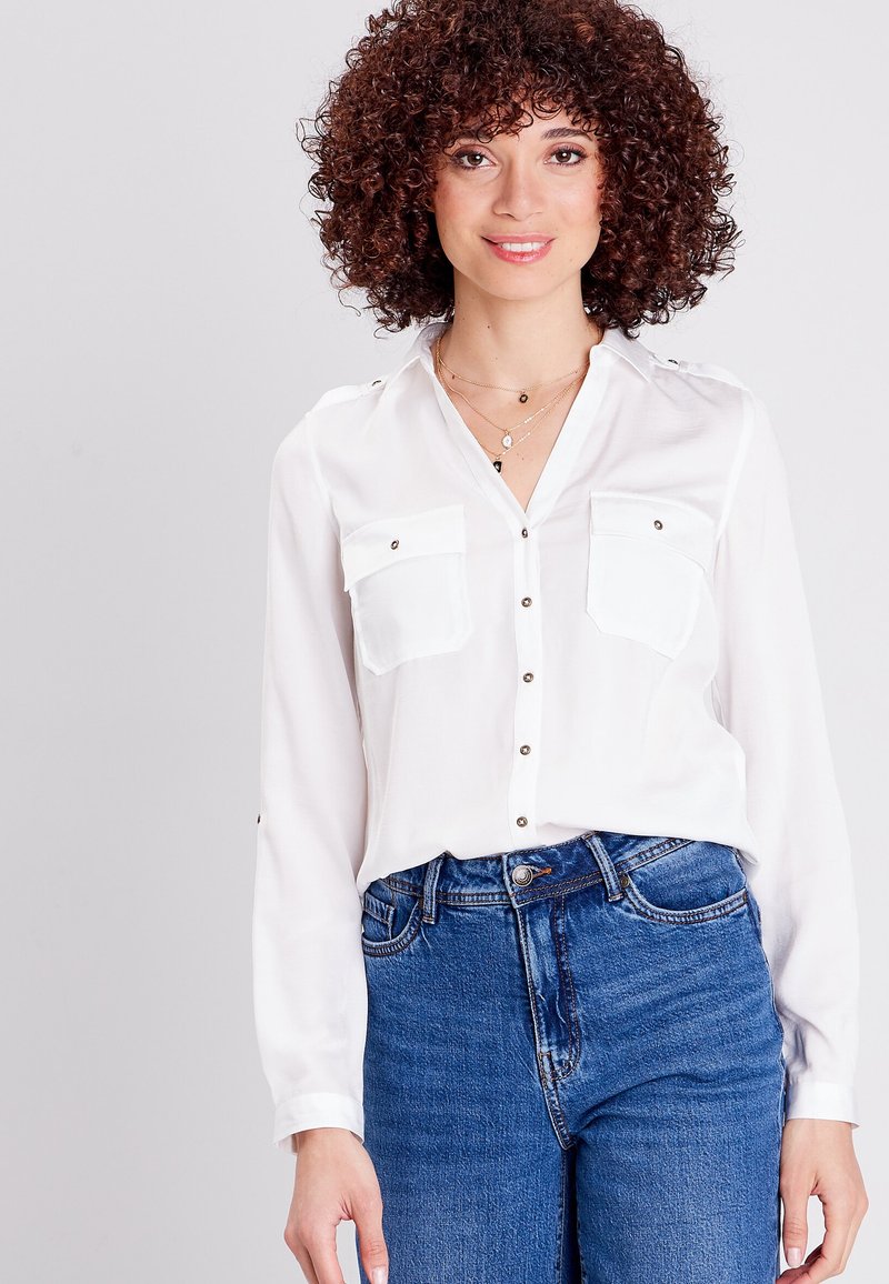 Blusa bianca a maniche lunghe con due tasche sul petto, chiusura frontale con bottoni e dettagli sulle spalle, abbinata a jeans blu a vita alta.