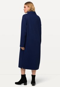 Langärmliges Kleid aus dunkelblauem Stoff mit geometrischem Muster, hochgeschlossen, mit seitlichem Schlitz und kombiniert mit knielangen schwarzen Stiefeln.