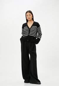 Schwarz-weiß gestreifter Cardigan aus weichem Strick mit V-Ausschnitt und Knopfverschluss. Kombiniert mit weit geschnittenen schwarzen Hosen.