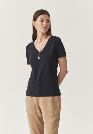Granatowa, krótko rękawowa koszulka typu V-neck z miękkiego materiału. W połączeniu z beżowymi spodniami. Widoczny złoty naszyjnik z wisiorkiem. Czysty design.