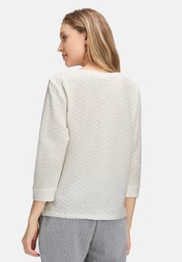 Femme aux longs cheveux blonds portant un haut blanc texturé à manches longues et un pantalon gris à rayures, de dos contre un fond blanc.