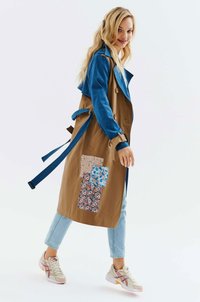 Płaszcz trench w brązowo-niebieskiej kolorystyce, ozdobiony łatami w kwiatowy wzór, z paskiem w talii i guzikami, zestawiony z jasnymi dżinsami i sneakersami.
