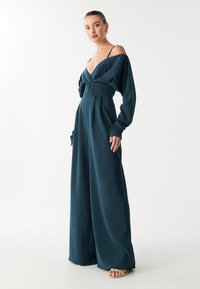 Tirkīza krāsas ilga piedurkņu kombinezons ar plecus atsedzošu dizainu, pieguļošu jostasvietu un plašiem, plūstošiem biksēm. Audums līdzīgs satīnam ar gludu tekstūru.