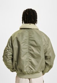 Person med mørke dreadlocks iført en oversized olivengrøn bomberjakke med lys fleecekrave, vender ryggen mod en ensfarvet baggrund.
