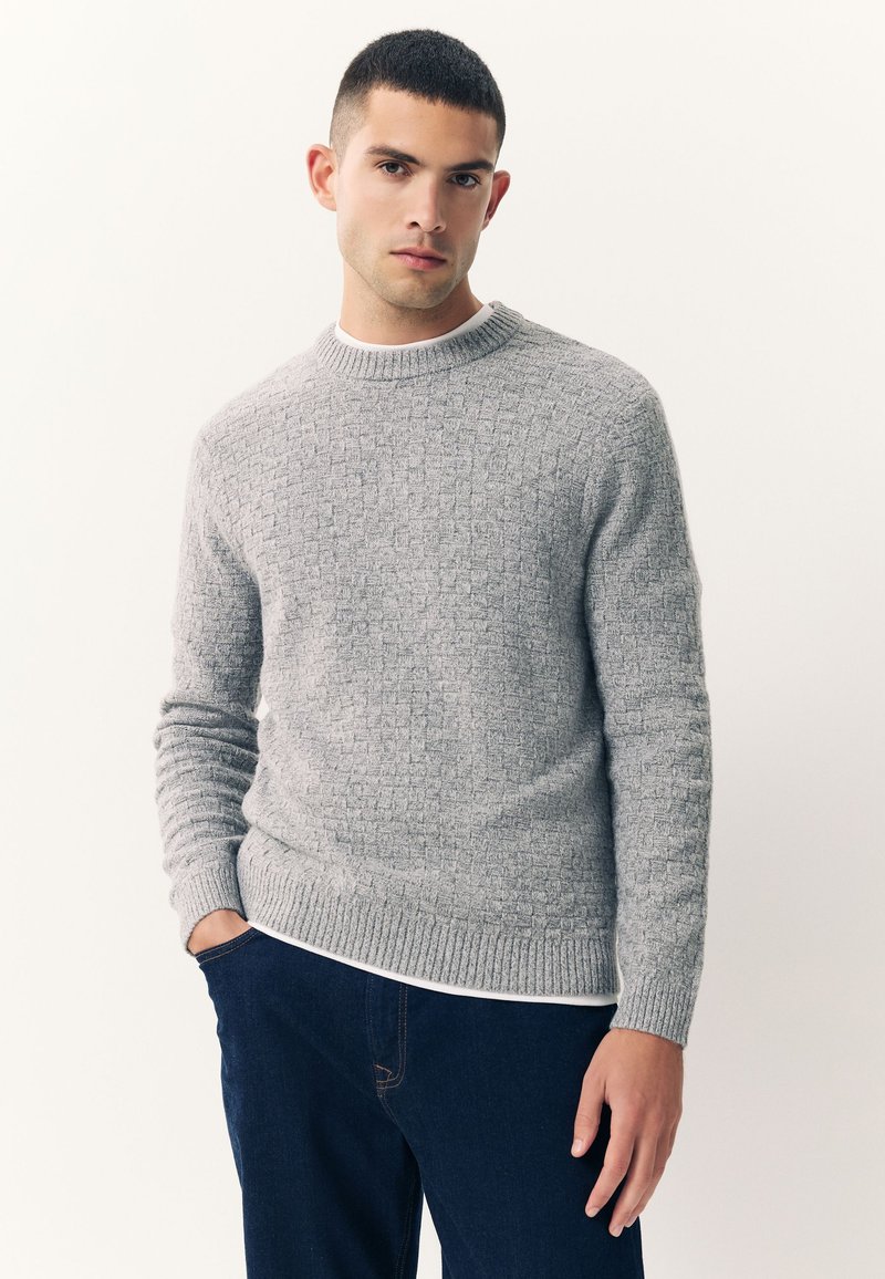 Maglione grigio a maglia testurizzata con scollo rotondo e polsini a costine, indossato sopra una maglietta bianca, abbinato a jeans blu scuro.