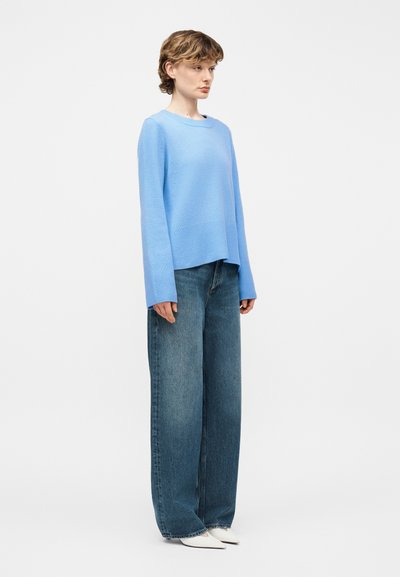 Νεαρός με κοντά σγουρά μαλλιά, φορώντας ένα ανοιχτό μπλε πουλόβερ, φαρδιά μπλε τζιν και λευκά μυτερά παπούτσια, στέκεται μπροστά σε ένα απλό φόντο.